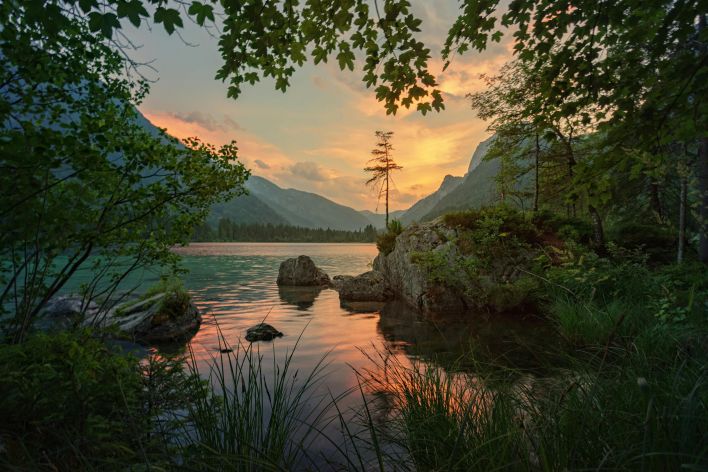 Peaceful sunset over a lake surrounded by mountains and lush greenery, capturing a tranquil natural scene.